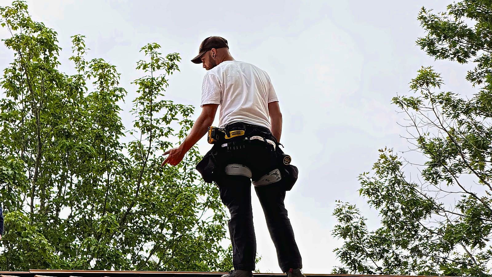 An installer in a tool belt standing on a roof, with trees behind