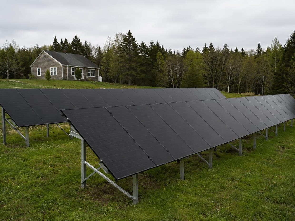 A cedar-shingle cottage backed by spruce trees, with two rows of ground-mount solar panels in the field